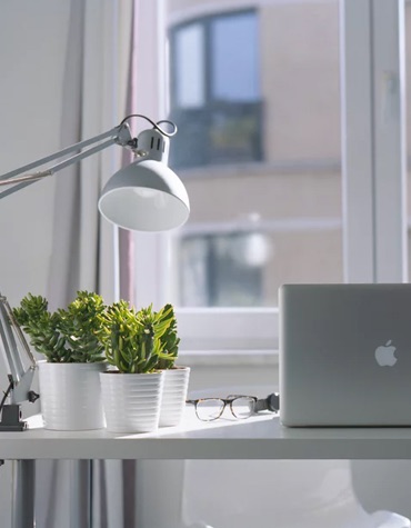 Three plants on a desk with a office lamp, glasses and macbook infront of a window