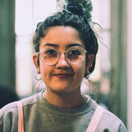 Woman with glasses wearing earrings, grey sweater and pink overalls